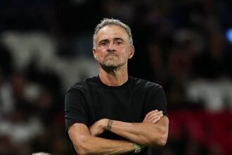 المدرب يقف مكتوف الذراعين مرتديًا قميصًا أسود في الملعب.